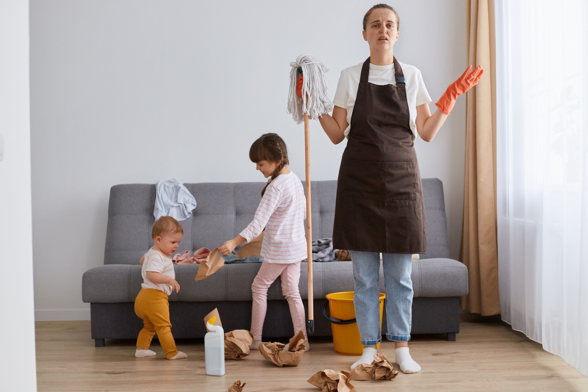 Indoor shot of young woman wearing white t shirt, brown apron and jeans, cleaning house, posing with her children, holding mop in hands, having helpless facial expression.